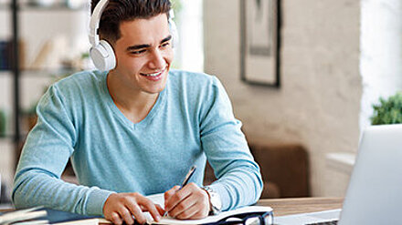 Bildung im Umbruch? Junger Mann sitzt aufmerksam lächelnd mit Kopfhörern vor seinem Laptop und macht sich Notizen.