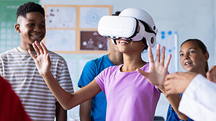 Lächelndes Mädchen mit VR-Brille bewegt sich mit ausgestreckten Händen durchs Klassenzimmer.