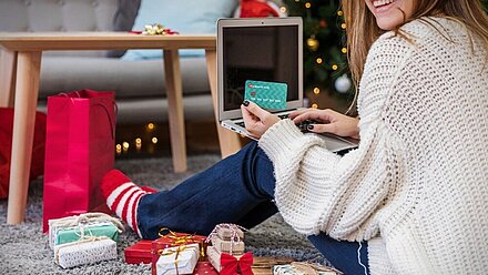 Compras navideñas: ¿qué buscan las familias españolas estas fiestas? Una mujer sonriente compra regalos navideños en línea, usando una tarjeta de crédito mientras está sentada en el suelo rodeada de paquetes envueltos.