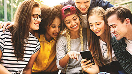 Gruppe Teenager sitzt zusammen und sieht interessiert lächelnd auf ein Smartphone.