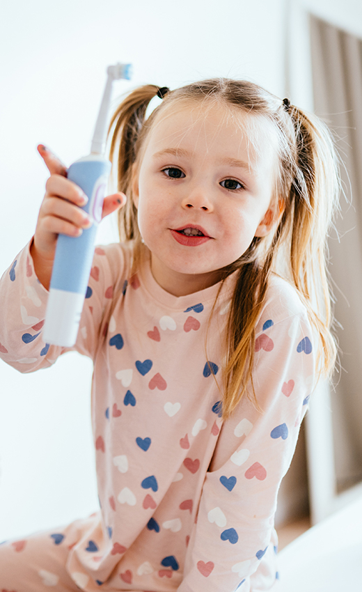 Kleines Mädchen in Pyjama hält eine elektrische Zahnbürste in der Hand.