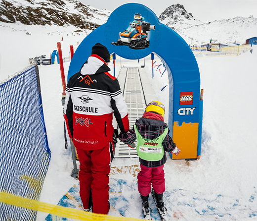 Eine erwachsene Person hält ein kleines Kind an der Hand. Sie tragen Skikleidung.