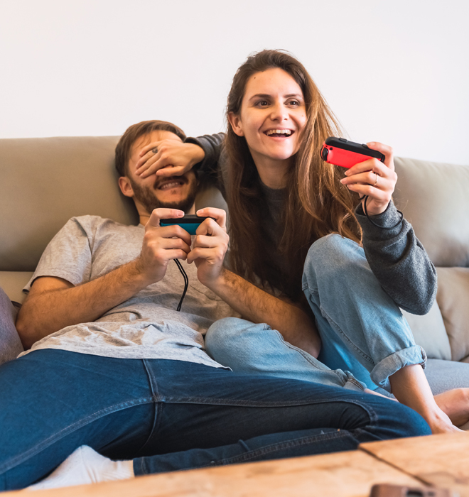 Junges Paar sitzt auf einem Sofa und hält einen Videocontroller in der Hand. Die Frau hält dem Mann die Augen zu.