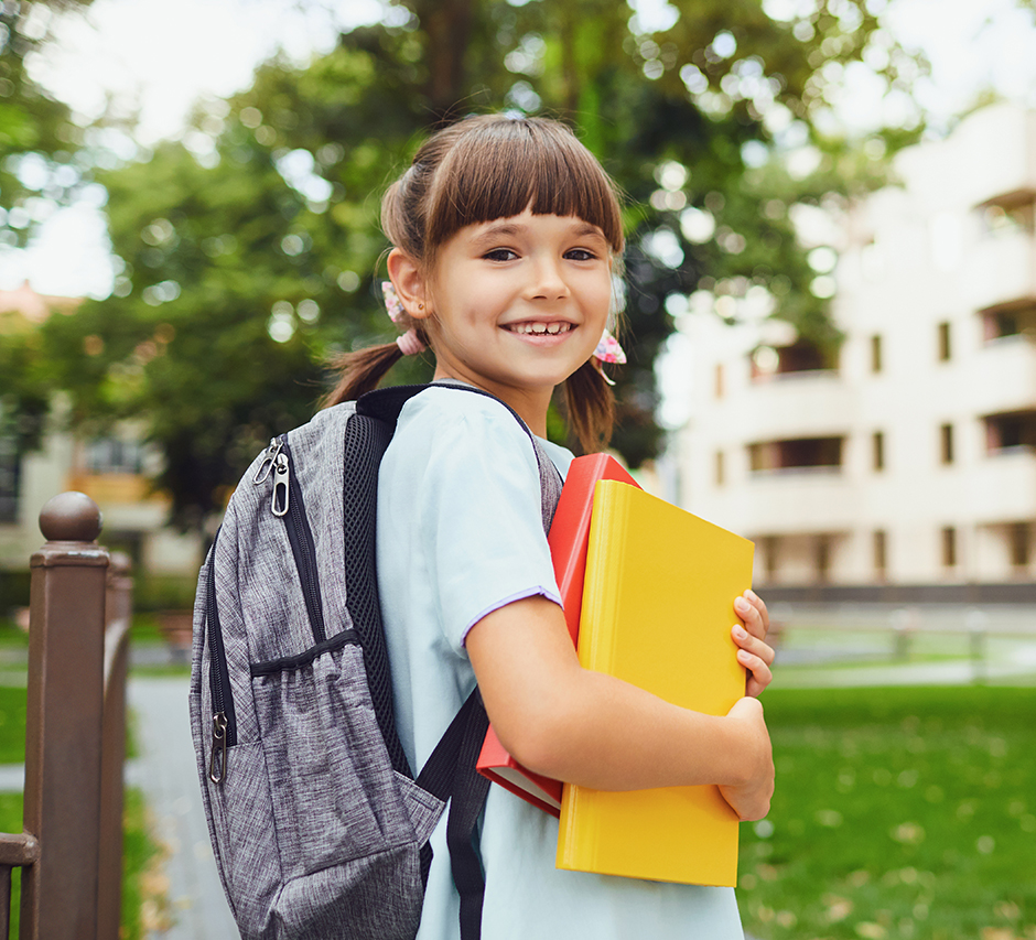 Kleines Mädchen mit dunklen Haaren und Rucksack hält ein gelbes Heft in der Hand und lächelt über die Schulter in die Kamera