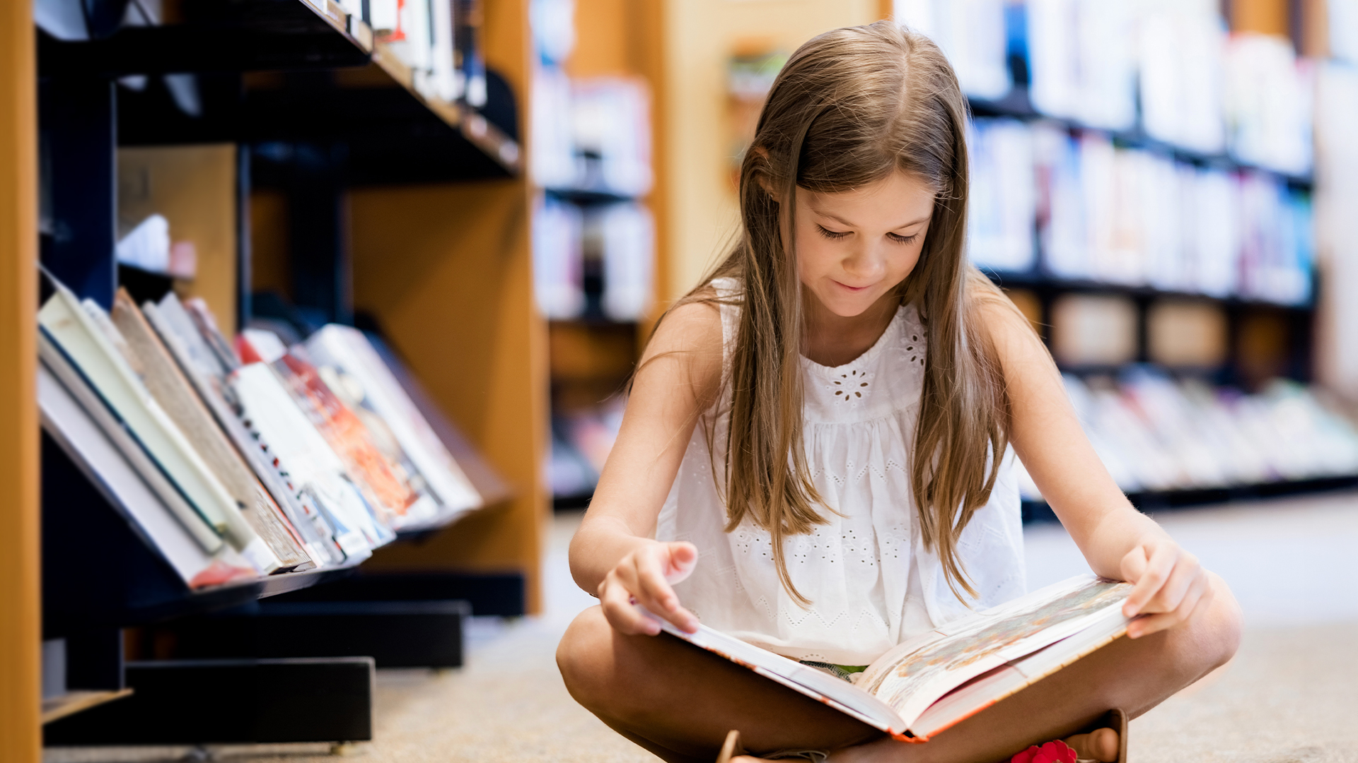 Junges Mädchen sitzt auf dem Boden zwischen Bücherregalen und liest ein Buch