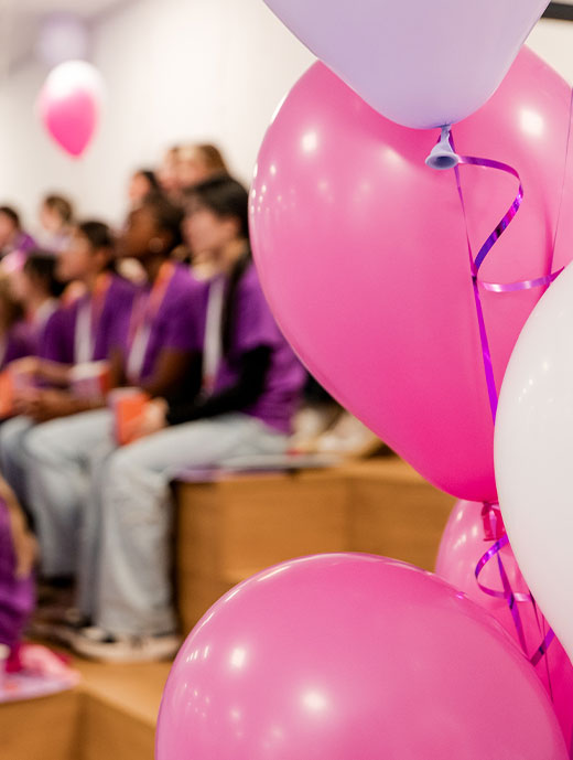 Pinke, weiße und lila Luftballons im Vordergrund und im Hintergrund verschwommen eine kleine Holztribühne auf der Menschen sitzen