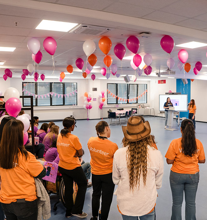 Gruppe Menschen mit orangenen Tshirts stehend von hinten. An der Decke sind orangene, pinke und weiße Luftballons