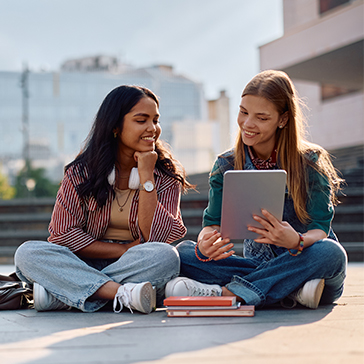 Zwei junge Frauen sitzen auf dem Boden draußen auf dem Boden und schauen gemeinsam in ein Tablet