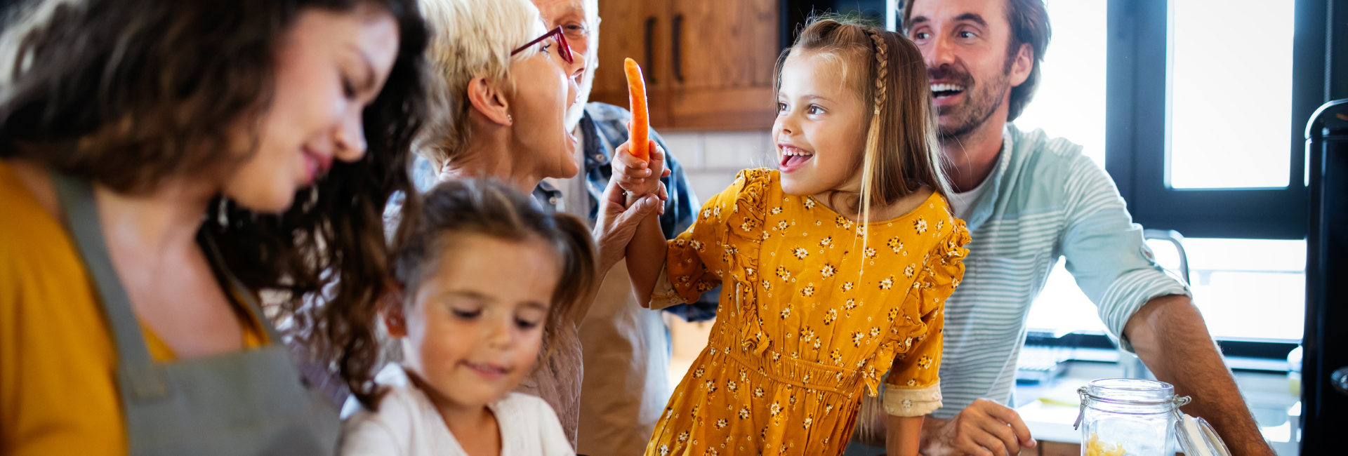 Eine Familie verbringt fröhlich Zeit miteinander in der Küche, wobei ein kleines Mädchen eine Karotte hält und ein älteres Familienmitglied anlächelt. Die Atmosphäre ist warm und voller Lachen.