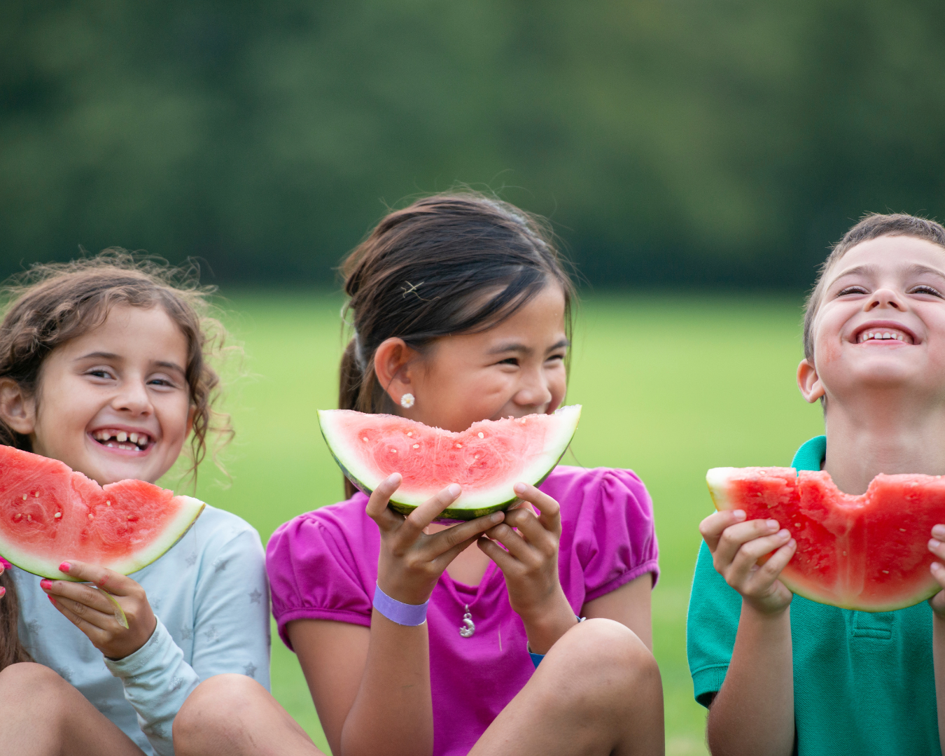 Tres niños sentados al aire libre, sonriendo y disfrutando de rebanadas de sandía en un día soleado, capturando un alegre momento de diversión veraniega.