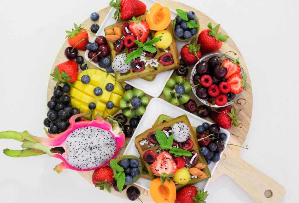 Un plato colorido lleno de frutas frescas como fresas, kiwis, moras, mangos y una pitahaya cortada. Las frutas están acompañadas de waffles decorados con frutas y hojas de menta.