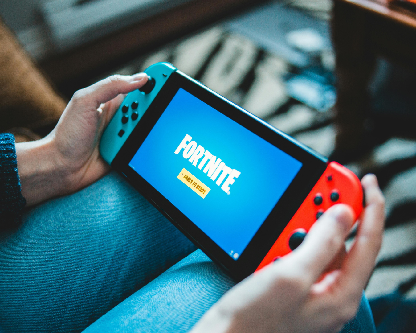 Persona sosteniendo una Nintendo Switch, mostrando la pantalla de título del juego «Fortnite» con la indicación «Pulsar para empezar.» La consola tiene mandos Joy-Con de color azul brillante y rojo.