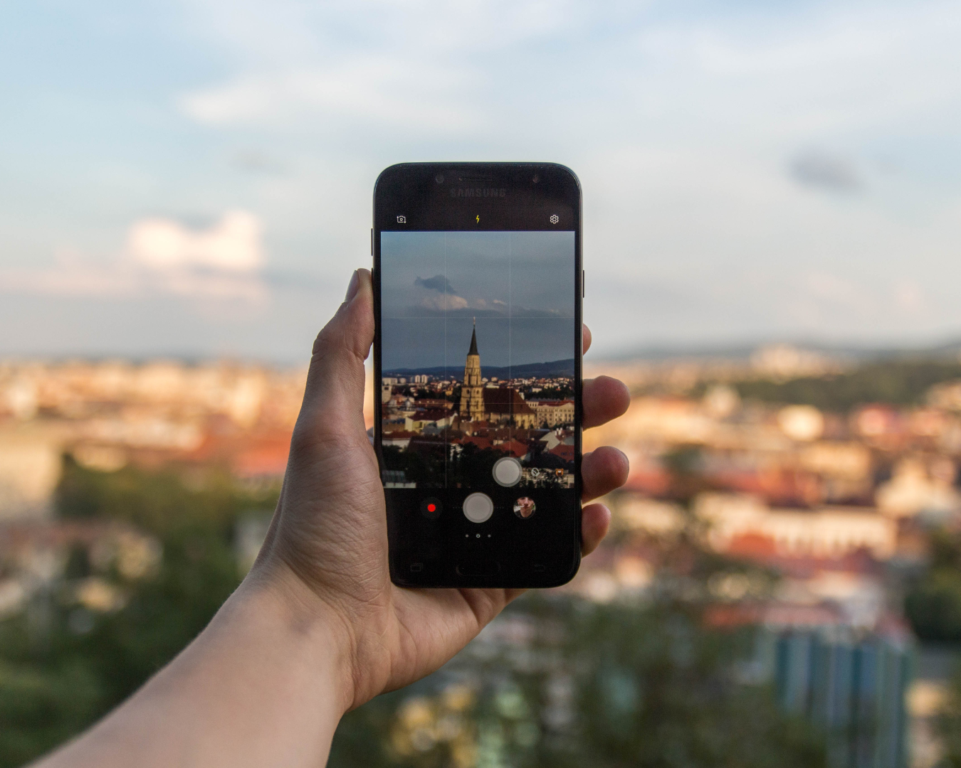 Una mano sostiene un teléfono capturando la vista de una ciudad al atardecer, combinando tecnología y paisaje en un momento tranquilo y contemplativo.
