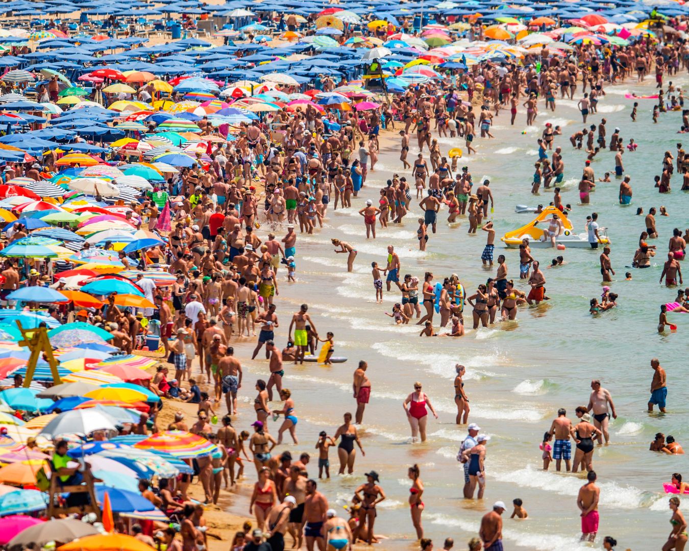 Una escena de playa abarrotada con numerosas personas en el agua y coloridas sombrillas cubriendo la arena, representando un ajetreado día de verano junto al mar.