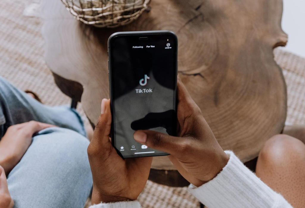 Una persona sostiene un smartphone y muestra la aplicación TikTok en la pantalla. El teléfono está enfocado, mientras que el fondo incluye una superficie de madera y la mano de la persona.