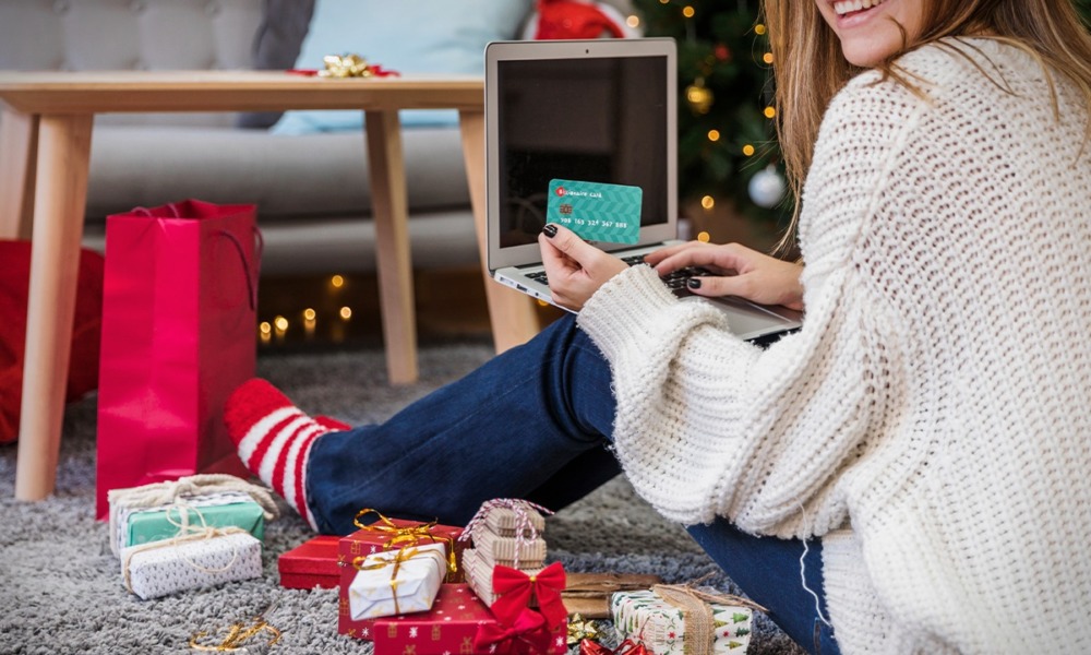 Una mujer sonriente compra regalos navideños en línea, usando una tarjeta de crédito mientras está sentada en el suelo rodeada de paquetes envueltos.