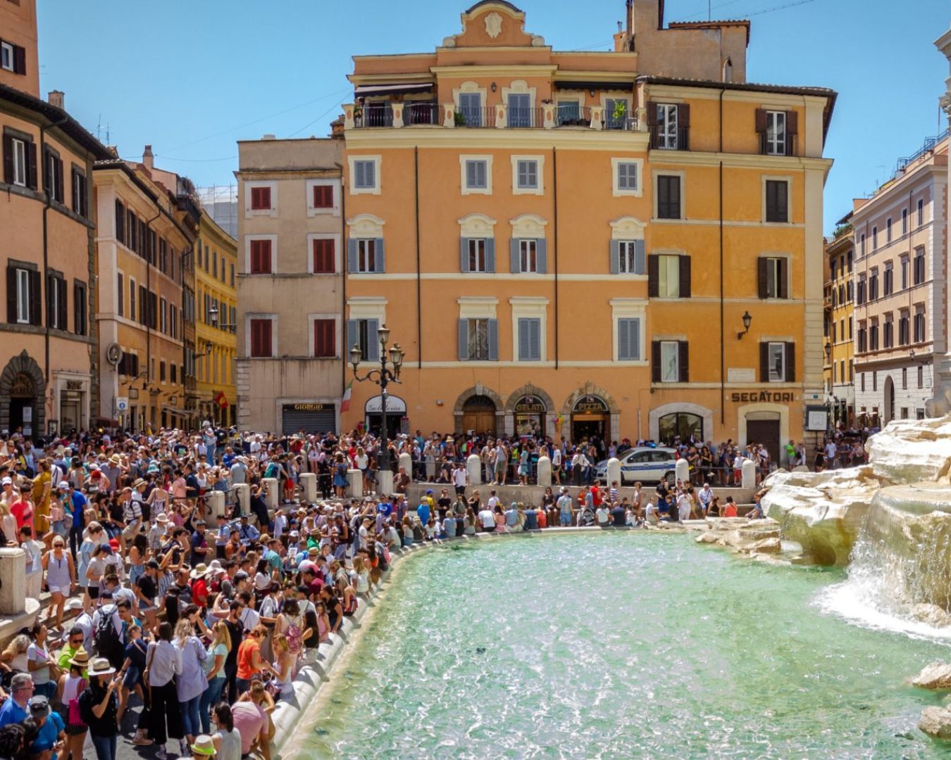 Una gran multitud se reúne alrededor de la Fontana de Trevi en Roma, con turistas llenando los alrededores en un día soleado. La escena pone de relieve la popularidad y la congestión de este famoso monumento.