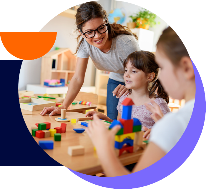 Two young girls are playing with building blocks at a table, guided by a smiling woman, likely a teacher, in a classroom setting. The scene emphasizes learning and creativity.