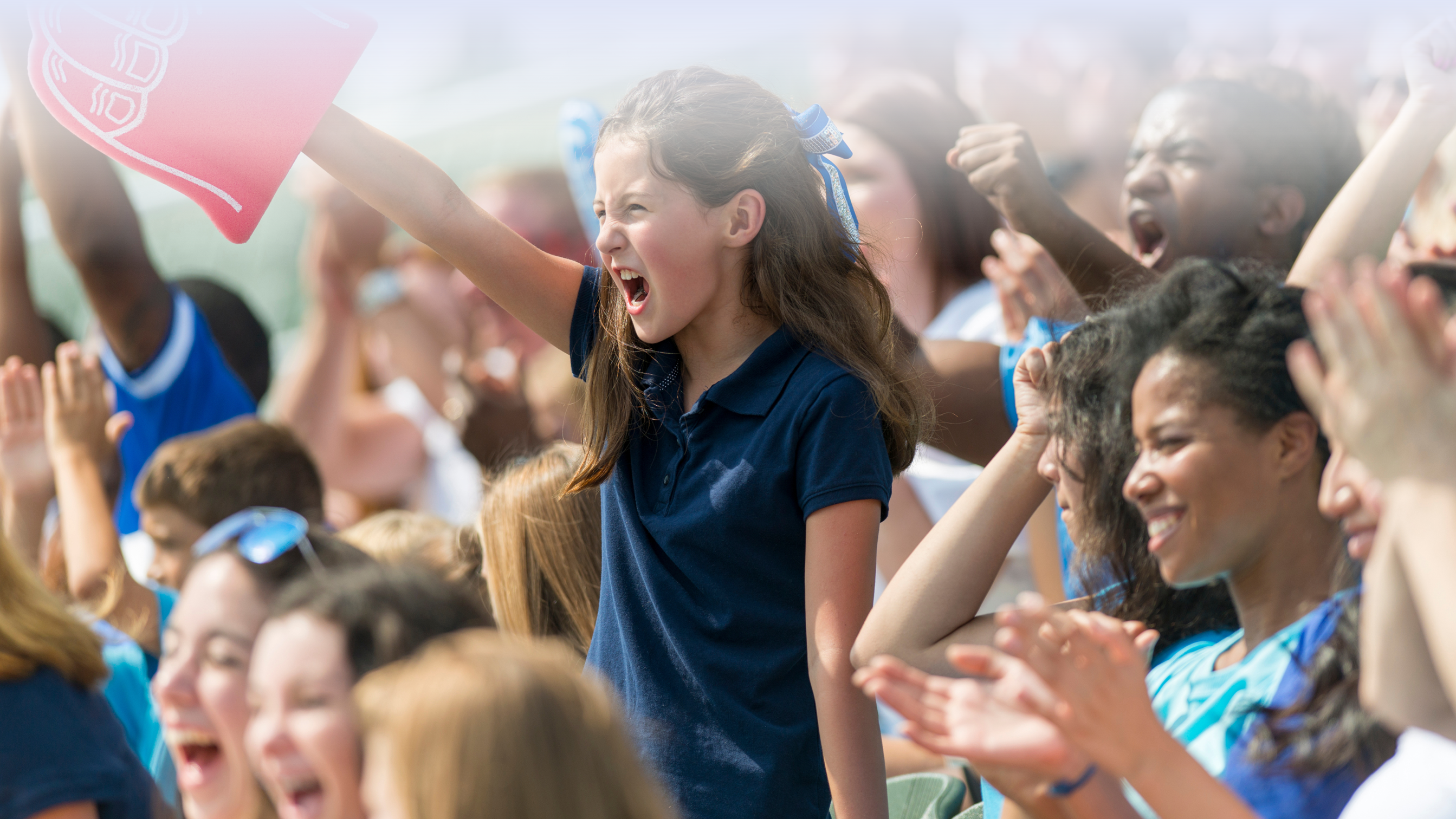 A group of people are cheering at a sports event. A girl in the centre stands in the foreground and is cheering on the team too.