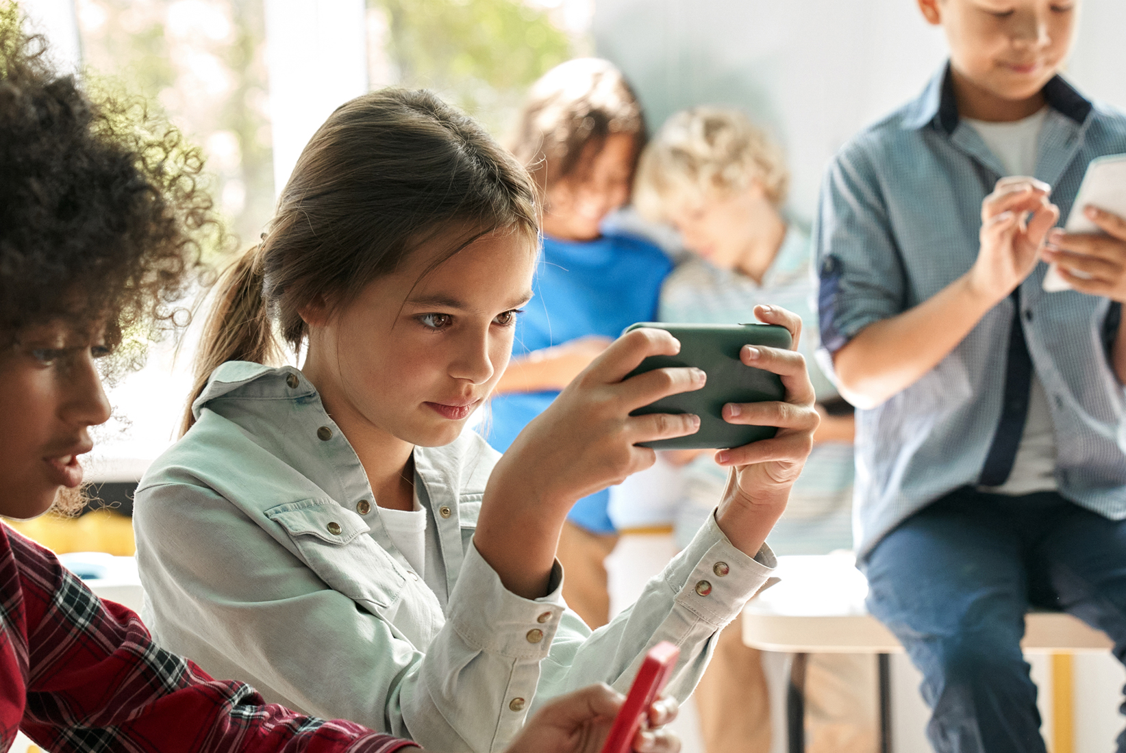 Young girl looking focused into a mobile phone and playing a game while other kids are around her.