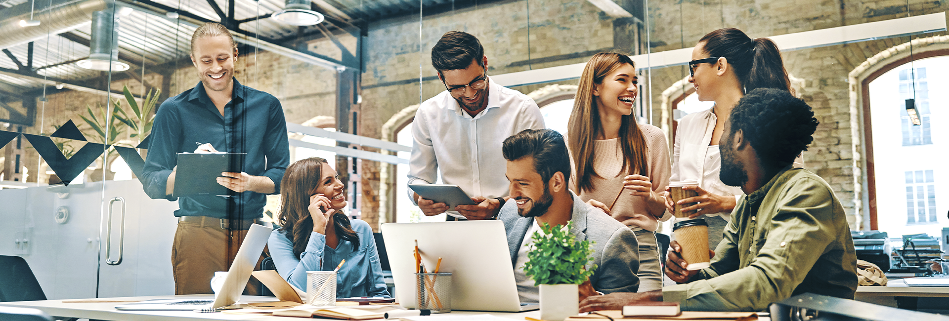 A group of people (men and women middle aged) are gathered around a table in the office with a laptop and a tablet smilingly looking at each other.