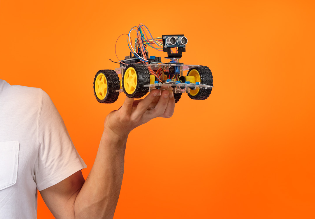 Small constructed car with visible cables is held in the hand of a boy. The background is orange.