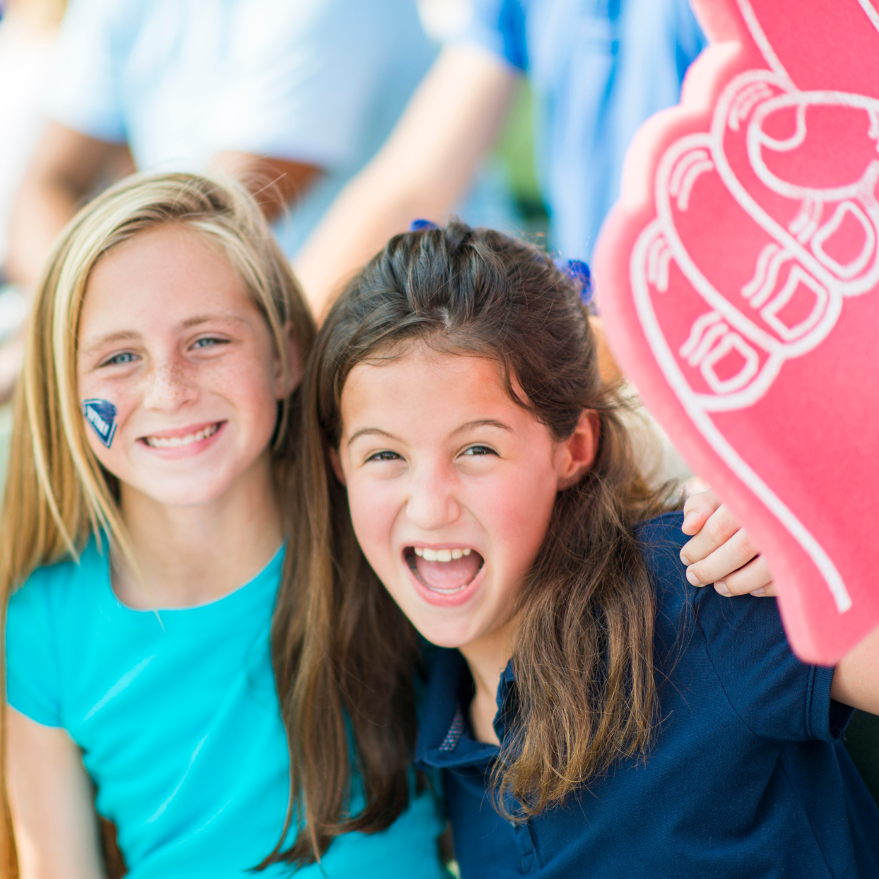 Two little girls look happily into the camera while laying their arms over each other. One girl is holding a large hand balloon with the index finger stretched upwards.