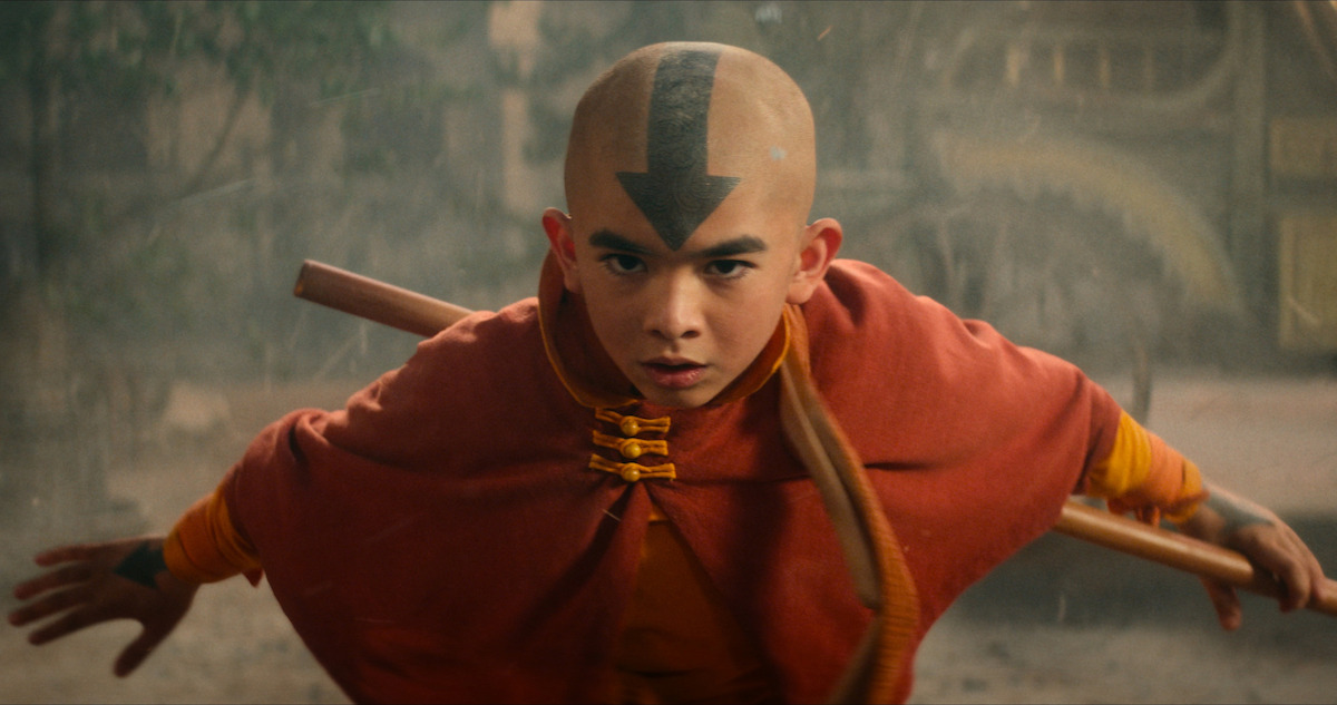 A young boy dressed as Aang from "Avatar: The Last Airbender," with the iconic arrow tattoo on his head, is in an action pose holding a staff, ready to fight.