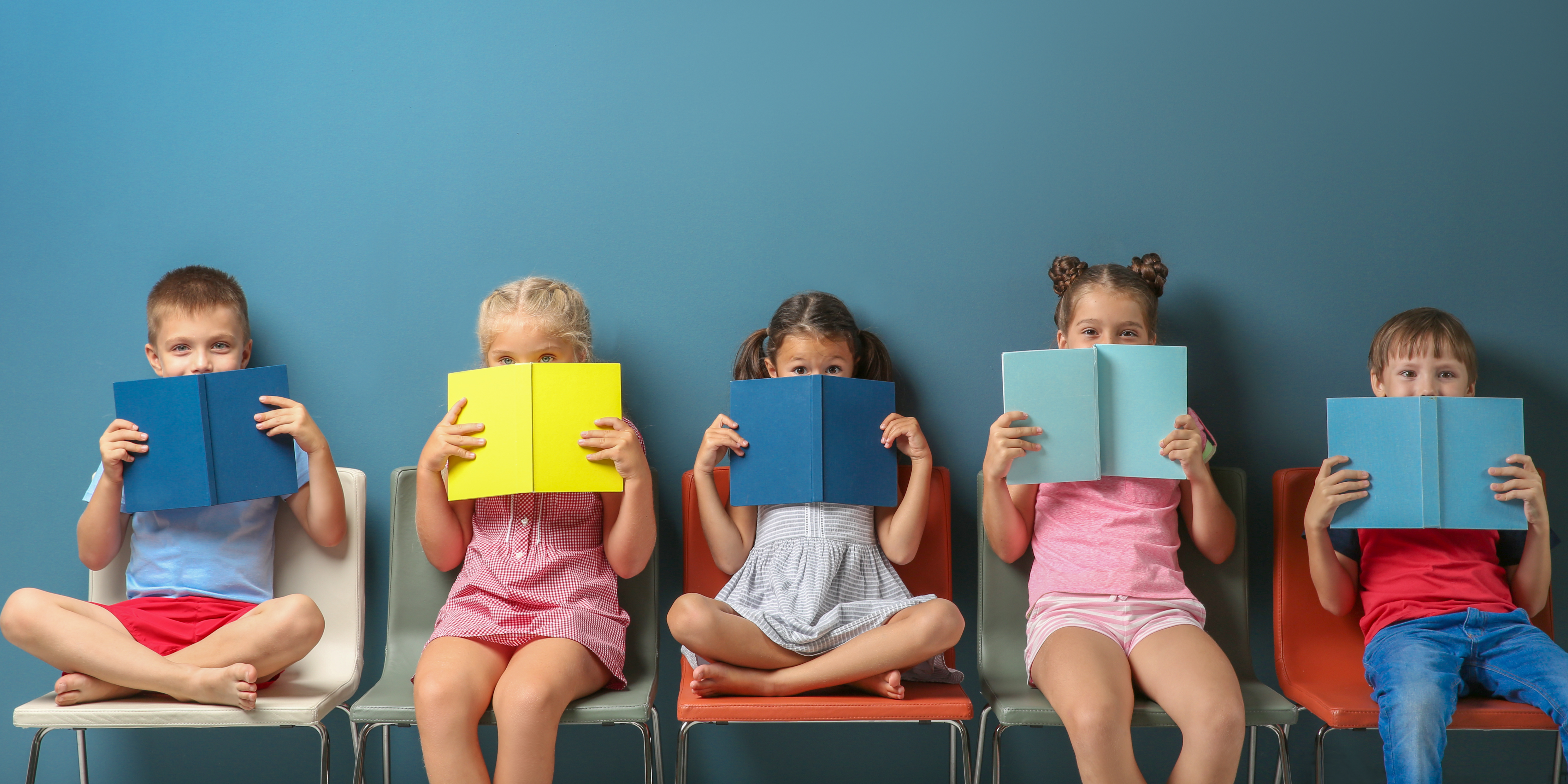 Young children sit on chairs, have a book open in front of them and smile at the camera.