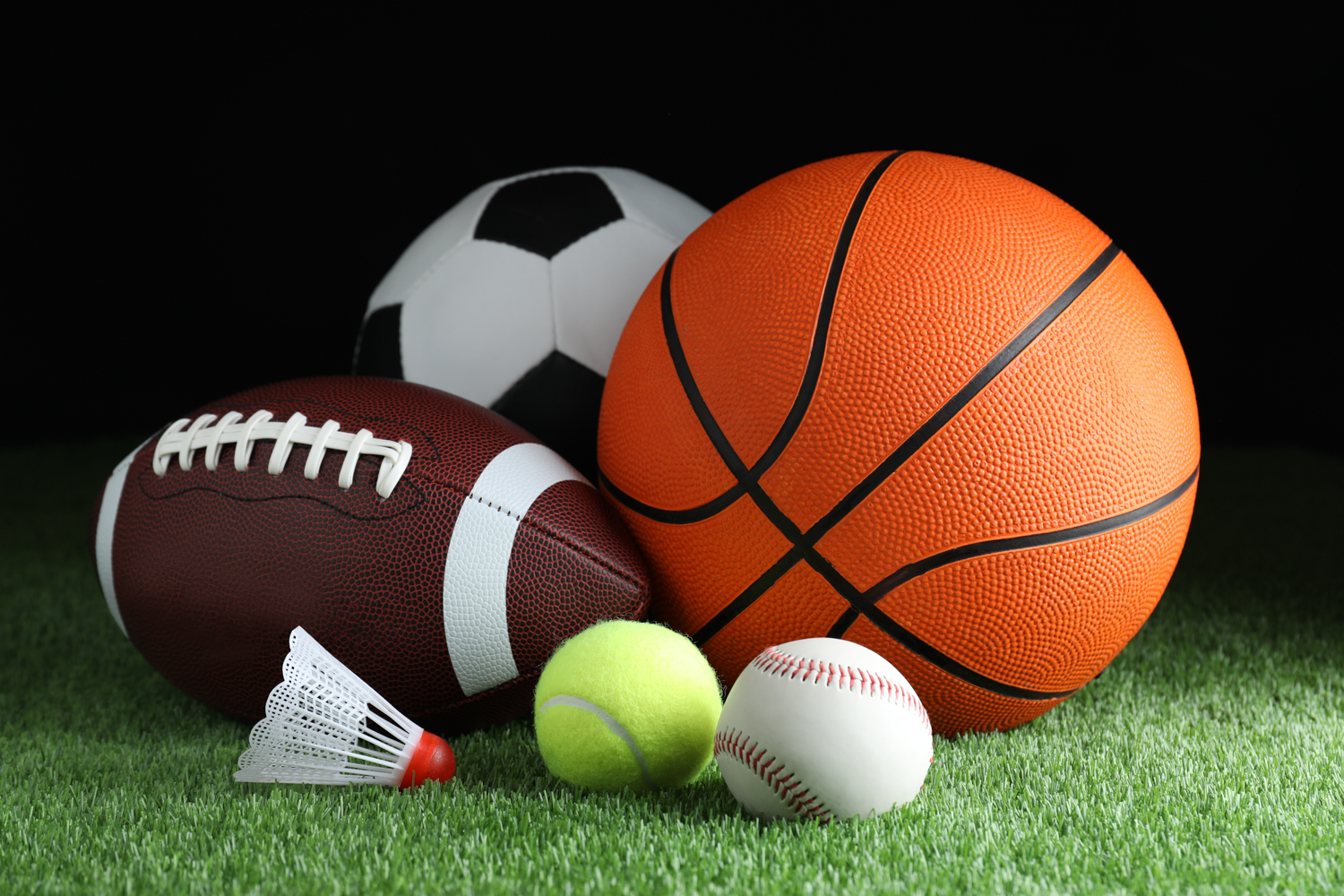 Several sports balls and a shuttlecock lie on green artificial turf against a black background.