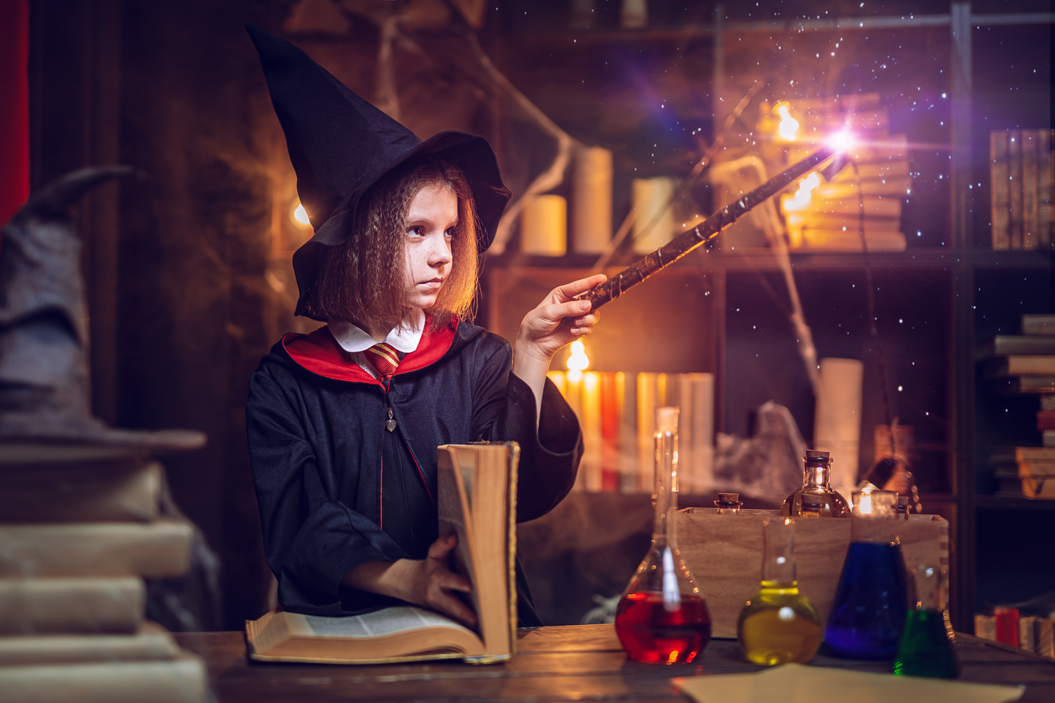 Child in a wizard costume pointing a wand at an open book in a magical setting. In the foreground are various colorful bottles and jars indicating alchemy or magic potions.
