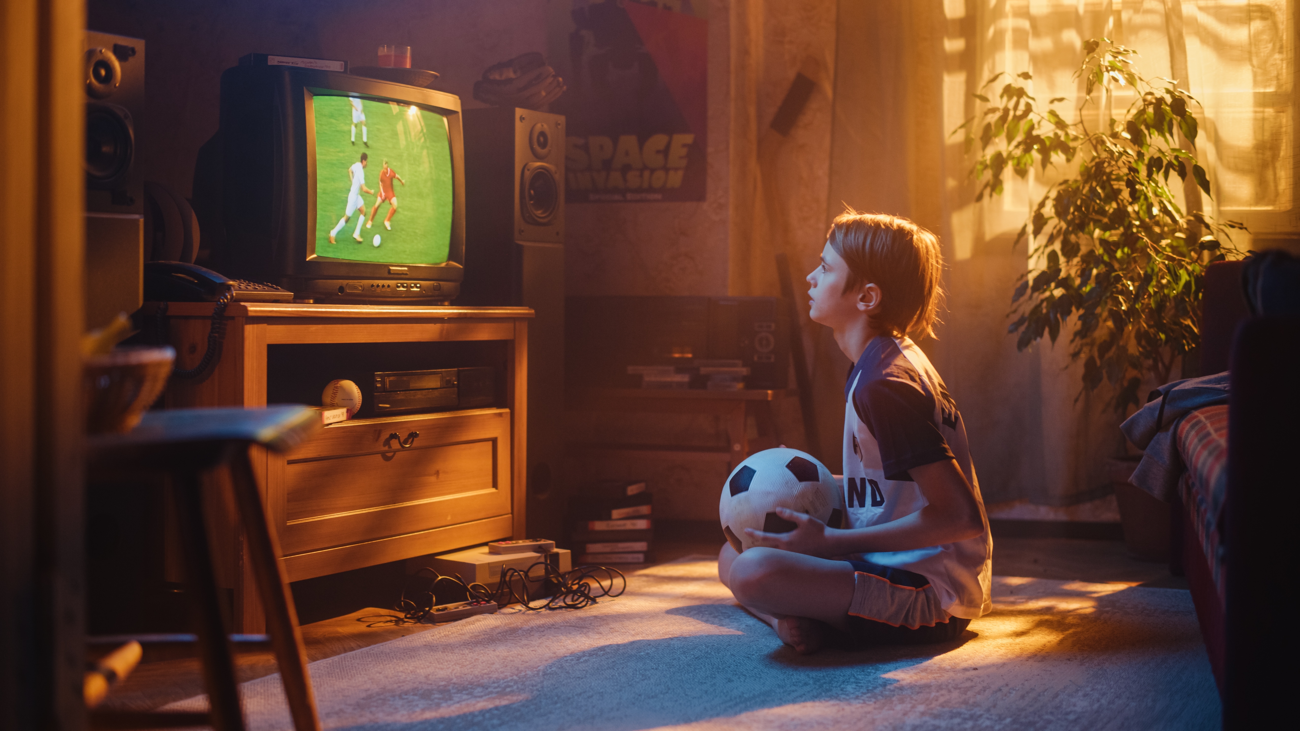 Boy sitting on the floor in the living room holding a soccer while watching a soccer match on TV.