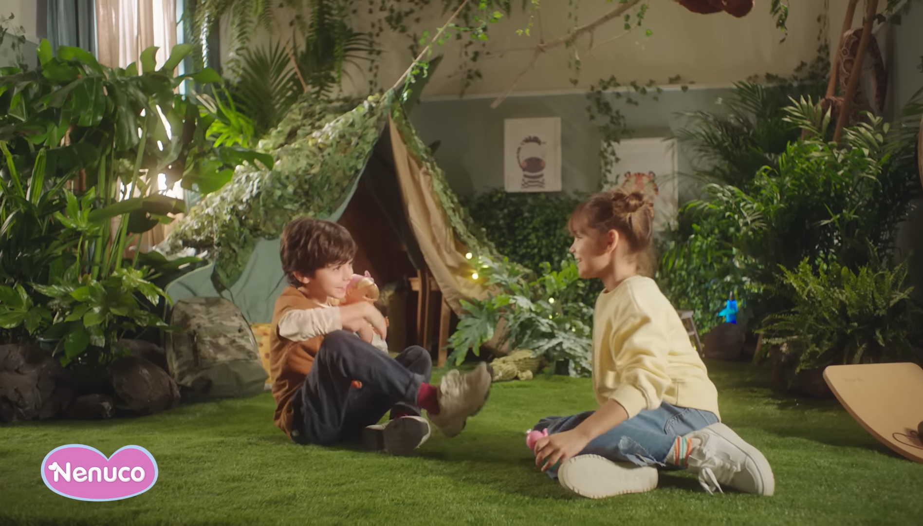 Girl and boy sit on an artificial meadow in front of an artificial "jungle" and play with Nenuco play dolls. They look happy and satisfied.