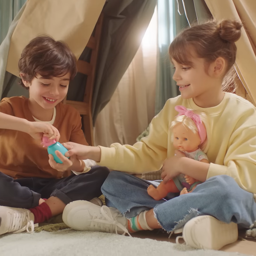 Girl and boy sit in a tent in the children's room and play with Nenuco play dolls. They look happy and content.