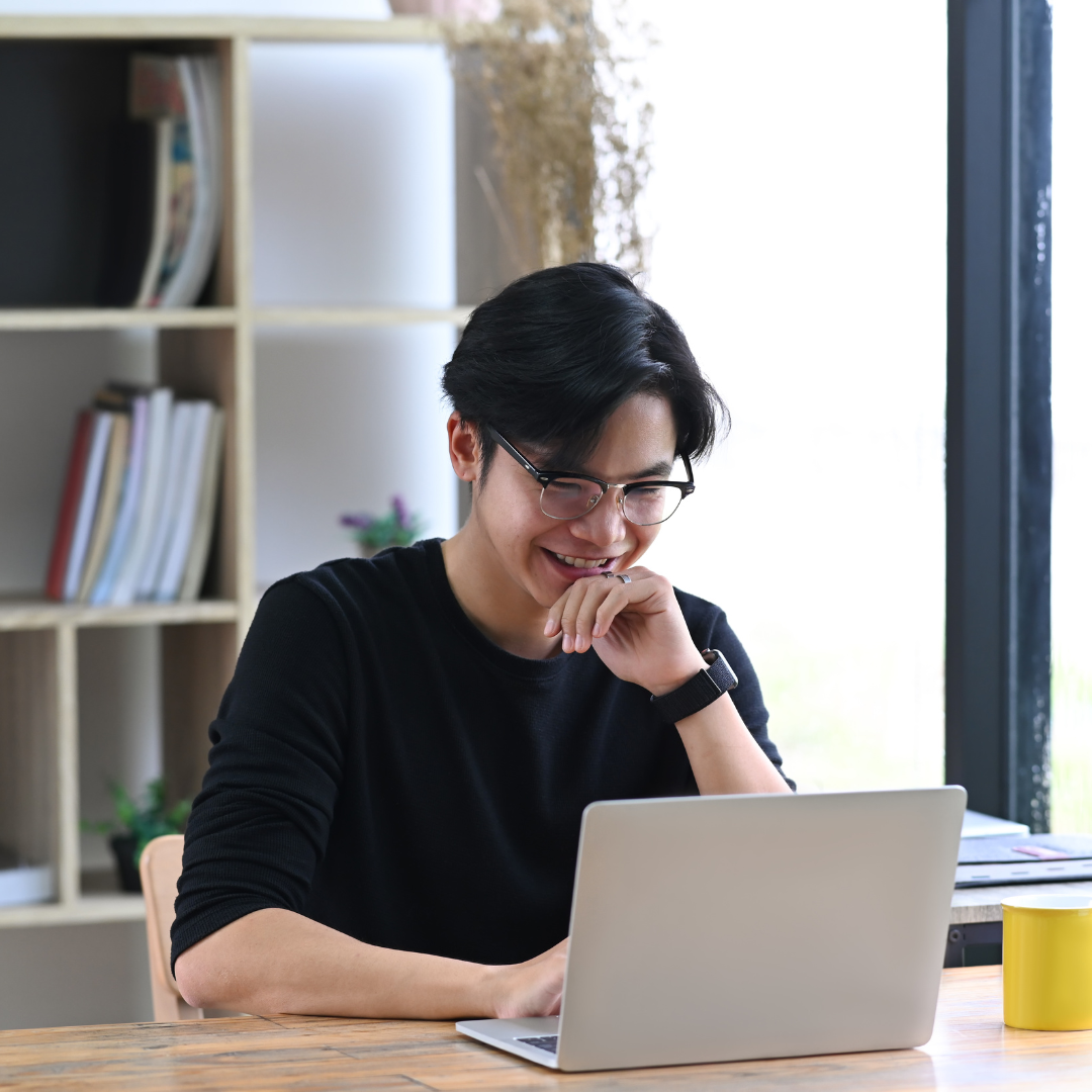 A young man sits at a table in front of his laptop and looks at the screen with a smile.