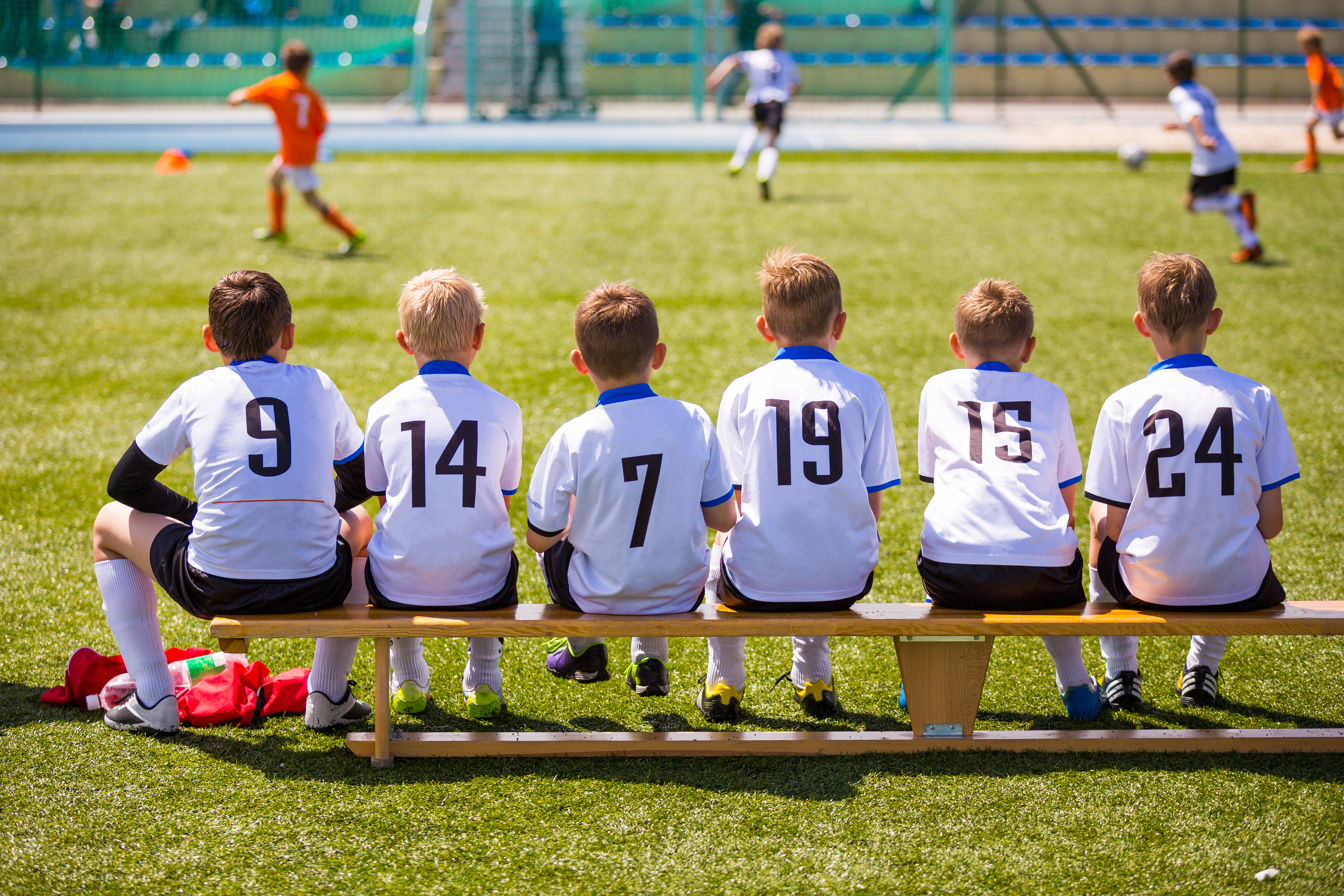 Young soccer players in white jerseys with numbers sit on a bench, watching teammates play on the field, showing team spirit and readiness to join the game.