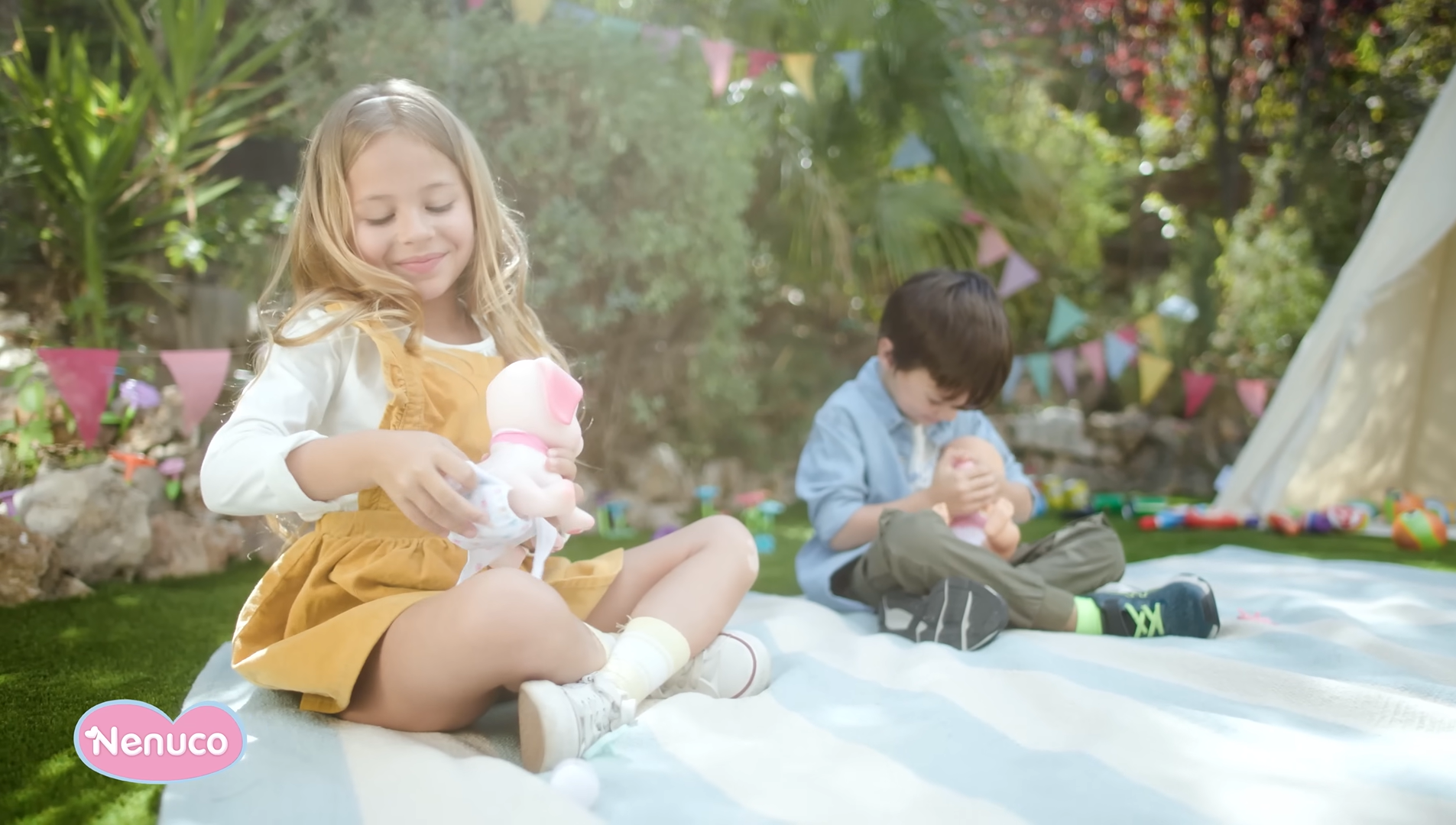 Girl and boy sit on a blanket in the garden and play with Nenuco play dolls. They look happy and satisfied.