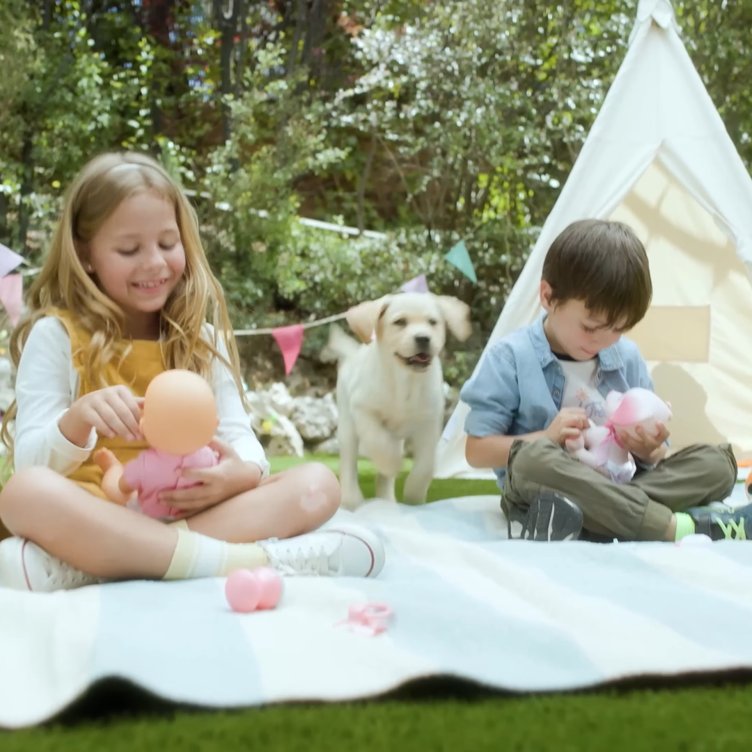 Girl and boy sit on a blanket in the garden in front of a tent and play with Nenuco play dolls. Behind them is a dog, who is also happy. They look happy and content.