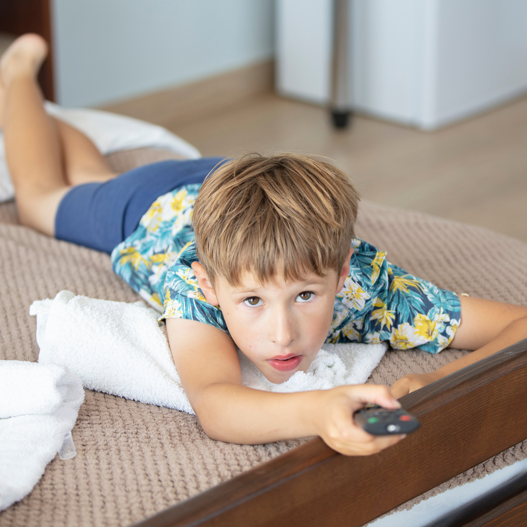 Little boy lying on the bed, holding a TV control and looking into the camera.