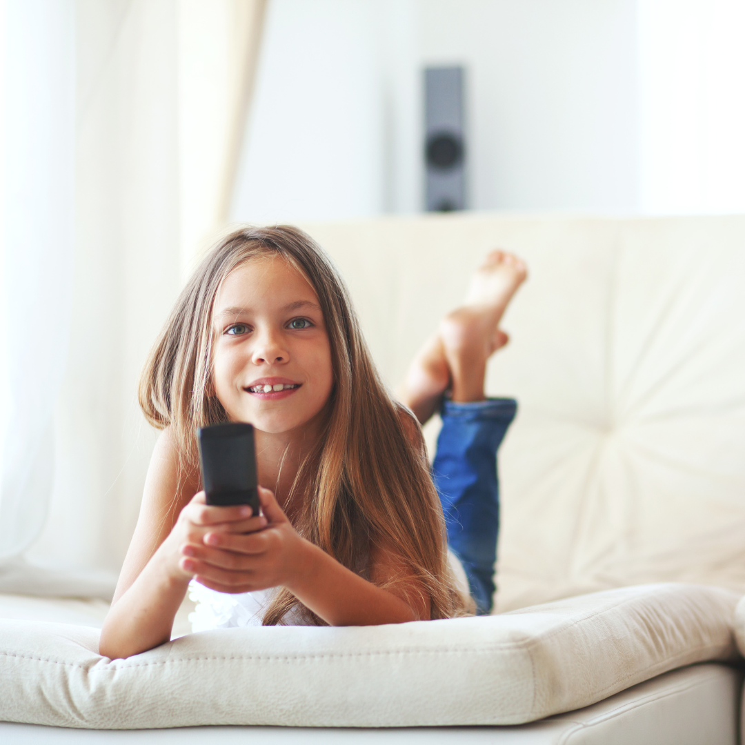 Little girl lying on the bed, holding a TV control in her hand and smiling as she looks towards the TV.