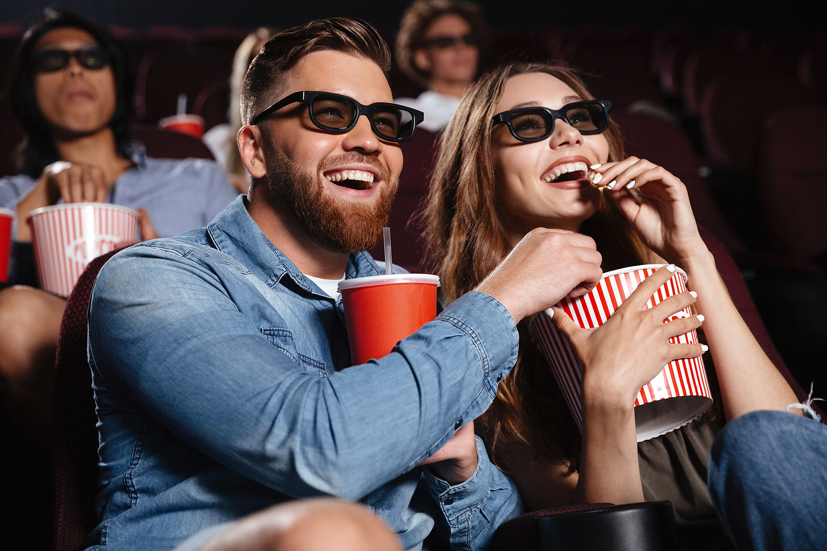 A woman and a man sit smiling in a cinema, watching a movie with 3D glasses and eating popcorn.